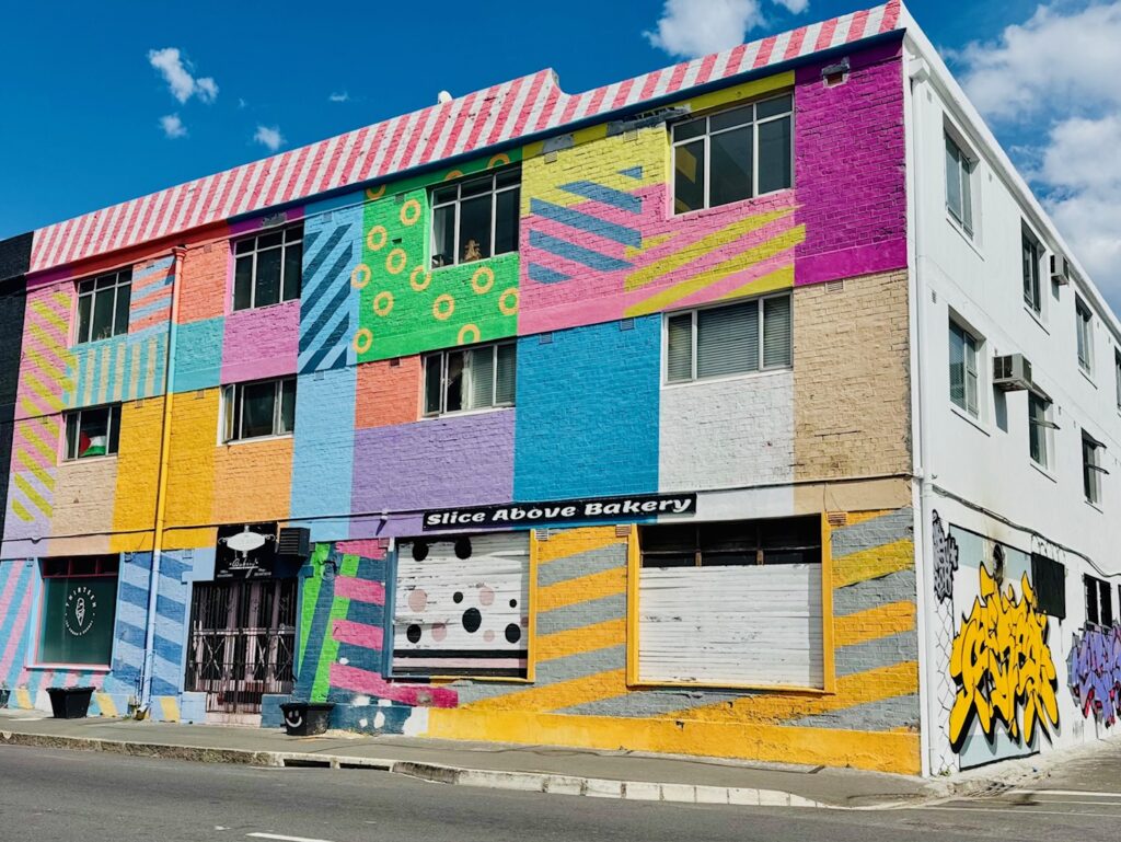 Mehrstöckiges Gebäude mit bunter, geometrischer und gestreifter Fassadenbemalung und Graffiti an der Seite