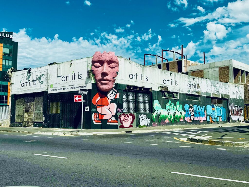 Eckgebäude mit Graffiti und einem großen, rosa Reliefgesicht sowie dem Schriftzug 'art it is' auf der Fassade unter blauem Himmel