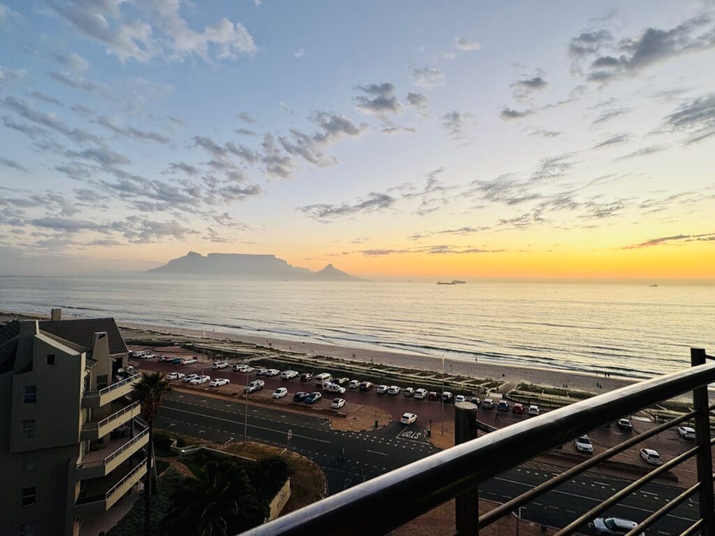 Blick von einem Balkon auf eine Küstenstraße mit parkenden Autos und dahinter das Meer sowie der Tafelberg in Kapstadt unter freundlichen Wolken bei Sonnenuntergang