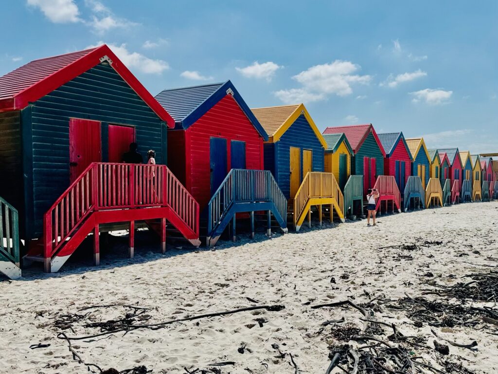 Reihe bunter Strandhäuser mit Treppen am Sandstrand von Muizenberg in Südafrika unter blauem Himmel