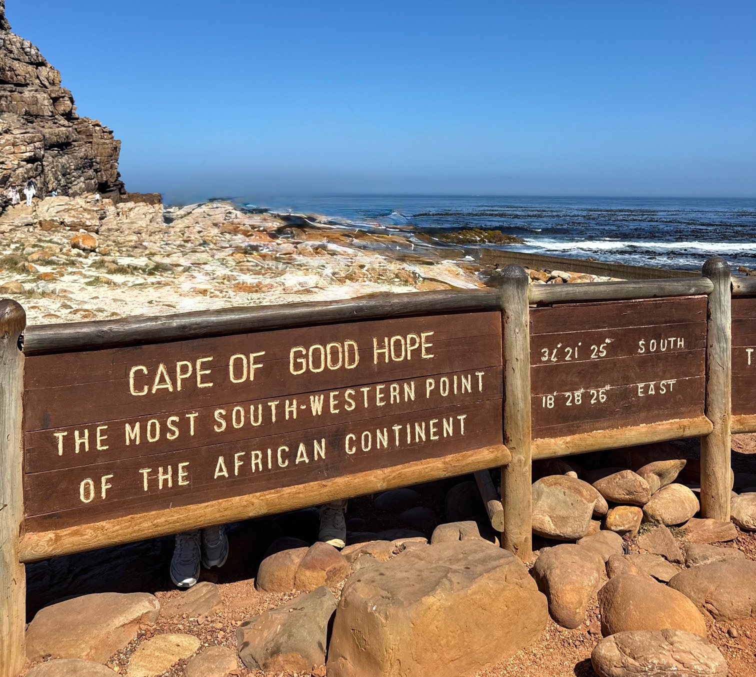 Holztafel mit der Aufschrift 'Cape of Good Hope the most south-western point of the African continent' vor felsiger Küstenlandschaft und Meer. Auf den Felsen unter der Tafel sind zwei Paar Turnschuhe zu sehen. Dieses Bild wurde mit KI bearbeitet: alle Personen, die an der Tafel posieren, wurden entfernt