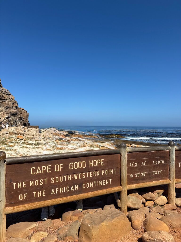 Holztafel mit der Aufschrift 'Cape of Good Hope the most south-western point of the African continent' vor felsiger Küstenlandschaft und Meer. Auf den Felsen unter der Tafel sind zwei Paar Turnschuhe zu sehen. Dieses Bild wurde mit KI bearbeitet: alle Personen, die an der Tafel posieren, wurden entfernt