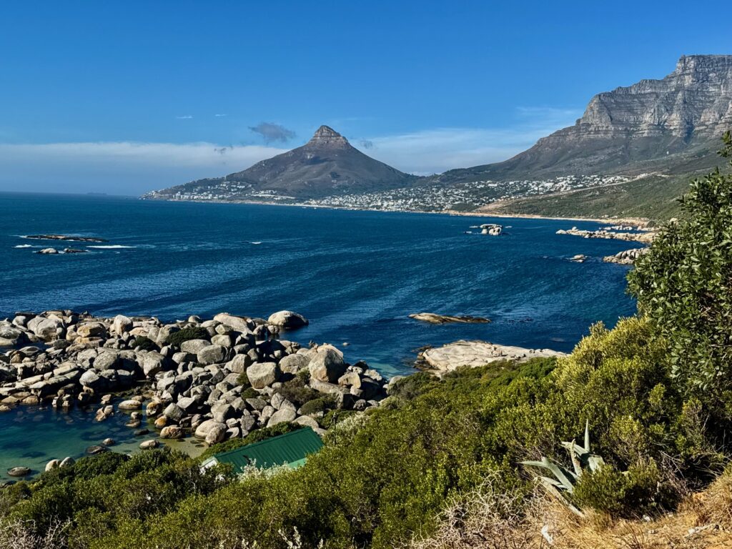 Bucht mit Felsen im Vordergrund, grünem Buschwerk und Bergen im Hintergrund unter blauem Himmel und mit tiefblau glitzerndem Meer