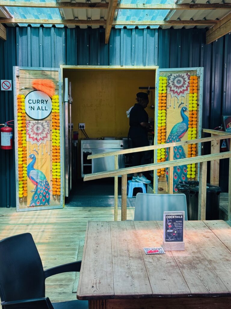 Offene Doppeltür mit bemalten Pfauen und Blumenketten, links ein Schild mit der Aufschrift 'CURRY 'N ALL', im Hintergrund eine Person in einer Küche, davor ein Holztisch mit Stuhl und Cocktailkarte