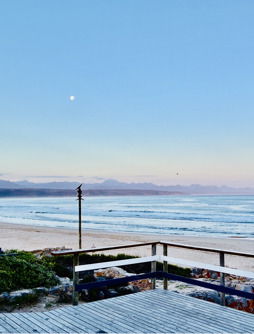 Blick von einer Holzterrasse mit Geländer auf einen Sandstrand und das Meer mit einer Bergkette unter klarem Himmel mit sichtbarem Mond