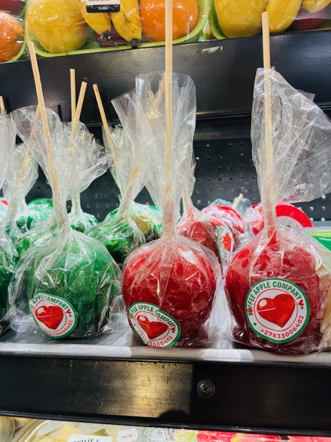 Mehrere grüne und rote kandierte Äpfel auf Holzstäbchen in durchsichtiger Folie verpackt mit Aufklebern der Marke The Apple Company. Im Hintergrund verpackte Bananen und Orangen.