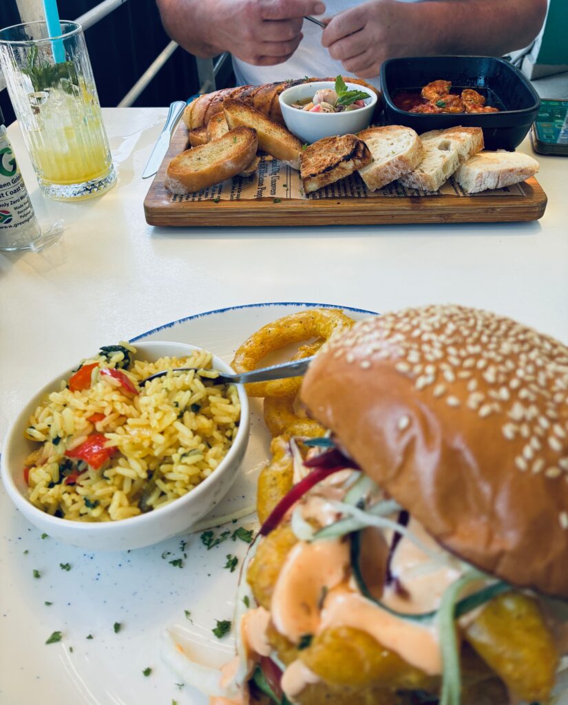 Nahaufnahme eines Burgers mit Sesambrötchen, Salat und Sauce, daneben eine Schale mit Reis und Gemüse sowie im Hintergrund ein Holzbrett mit verschiedenen Brotscheiben und Dips