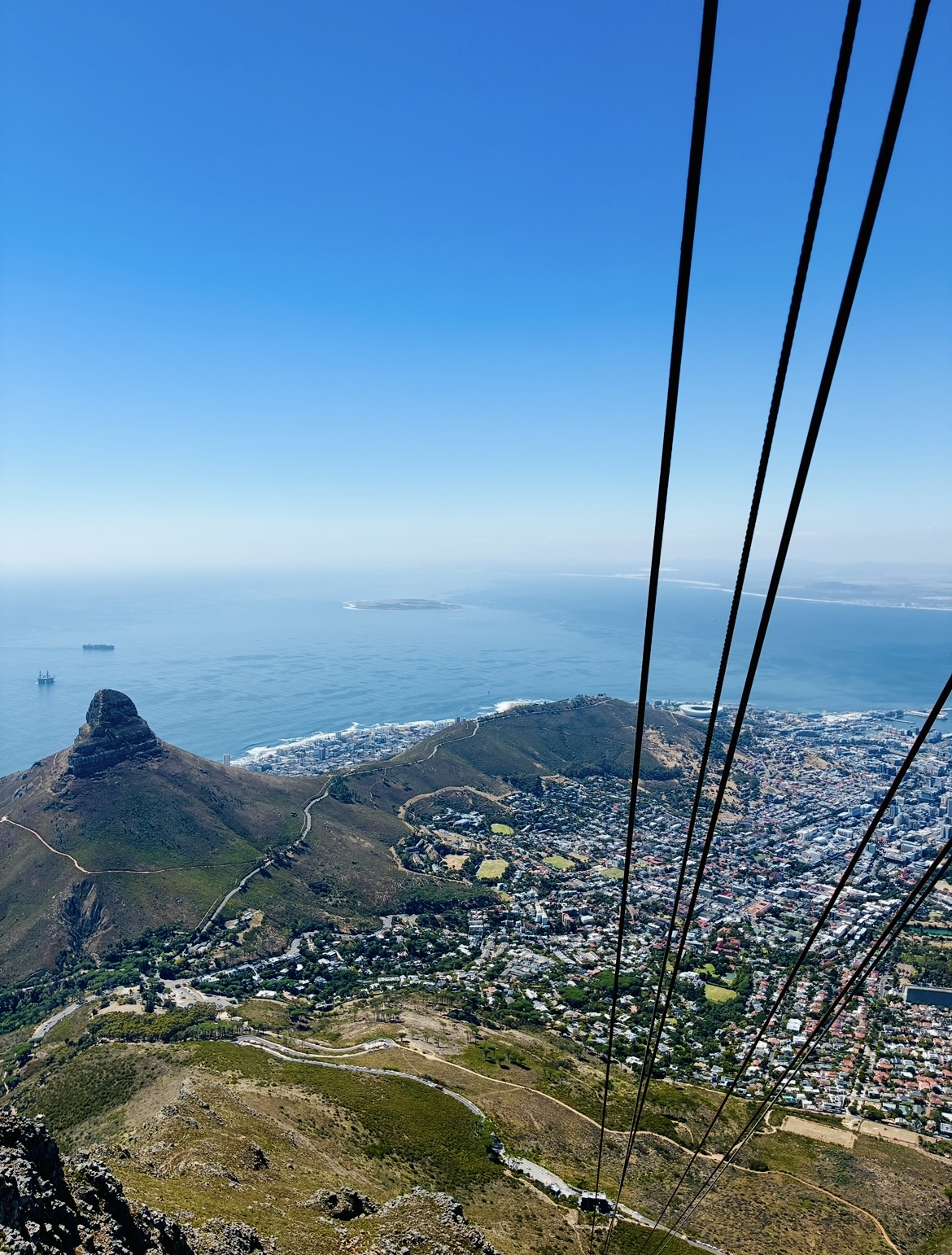 Blick von der Seilbahn zum Tafelberg mit Seilbahnkabeln auf Kapstadt mit Meer und mehreren Schiffen im Hintergrund