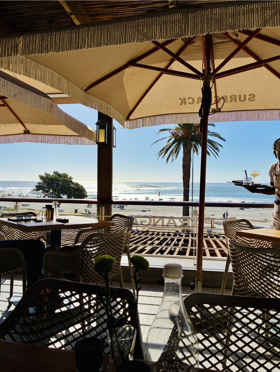 Blick von einem Café mit Sonnenschirmen und Flechtstühlen auf einen Strand mit Palmen und Meer im Hintergrund