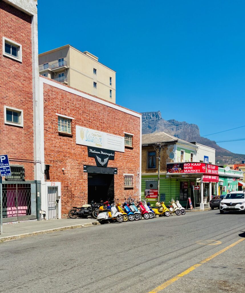 Straßenszene mit mehreren in Reih und Glied geparkten Rollern vor einem roten Backsteingebäude und einem Eckgeschäft an einer sonnigen Kreuzung mit dem Tafelberg in Kapstadt im Hintergrund
