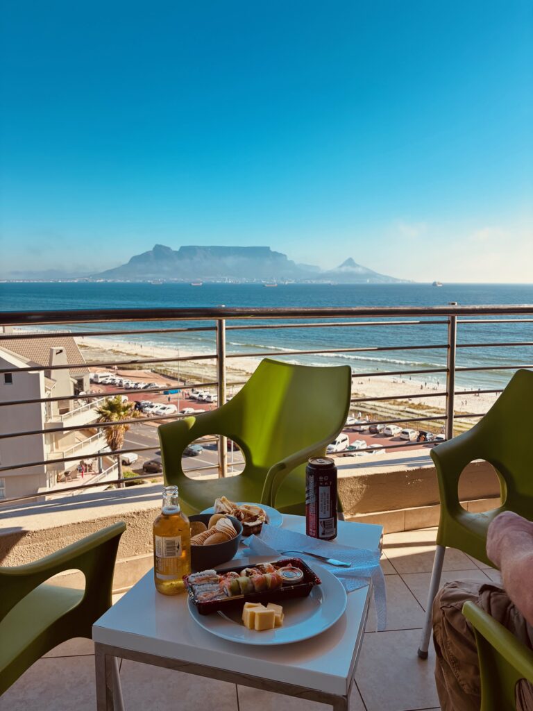 Balkon mit grünem Stuhl, weißem Tisch mit Sushi, Getränken und Käse, im Hintergrund Meer und Tafelberg unter blauem Himmel