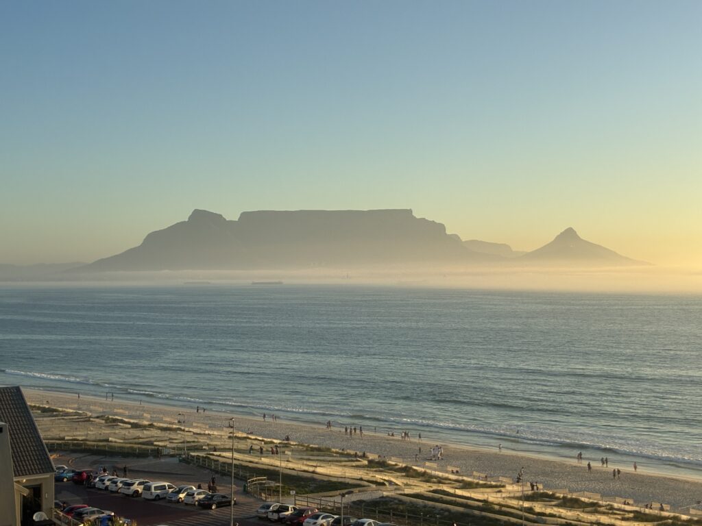 Blick auf einen Strand mit Menschen und Autos im Vordergrund und dem Tafelberg sowie dem Lion`s Head in Kapstadt im Hintergrund unter klarem Himmel mit leichtem Dunst
