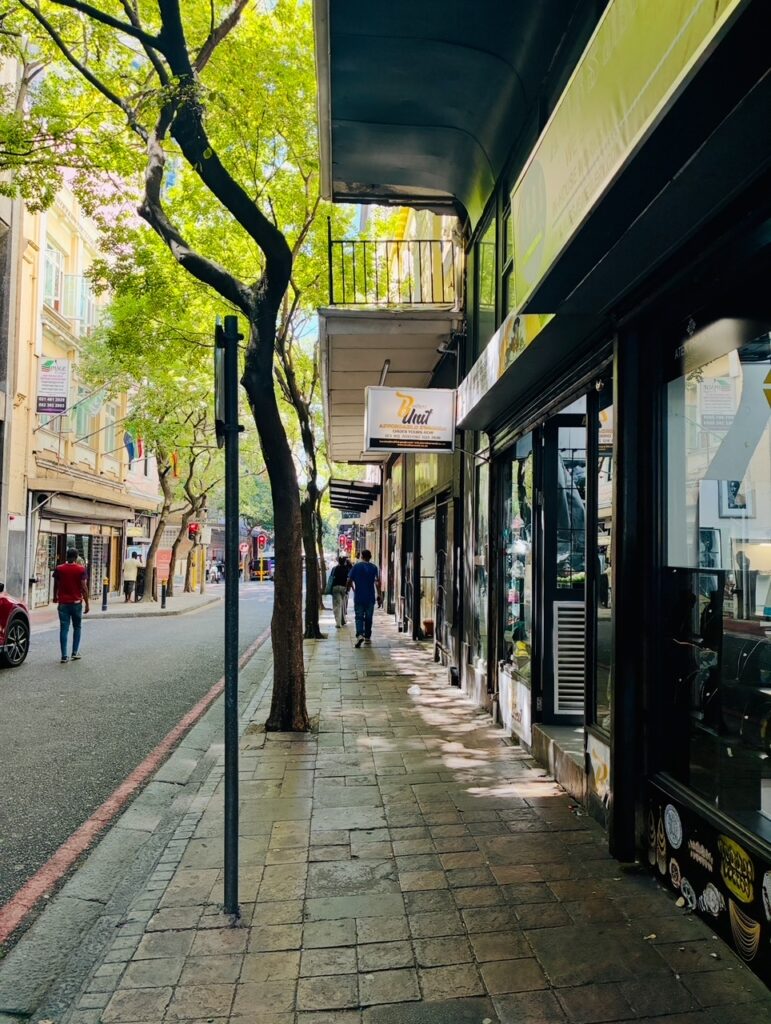 Straßenszene mit Gehweg, Bäumen, mehreren Passanten und Geschäften mit Schaufenstern