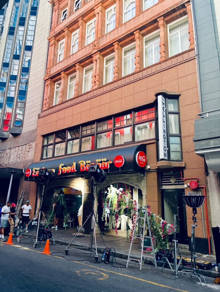 Straßenszene mit einem mehrstöckigen Gebäude, dessen Erdgeschoss einen Eingang mit dem Schriftzug 'Food Bazaar' und 'Lexington' zeigt, davor stehen Beleuchtungsausrüstung und Absperrkegel für Filmdreharbeiten