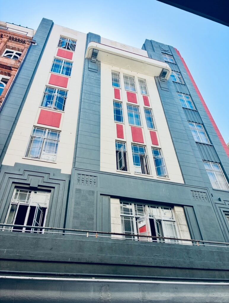 Fassade eines mehrstöckigen, vom Jugendstil beeinflussten Gebäudes mit weißen, grauen und roten Flächen sowie zahlreichen Fenstern