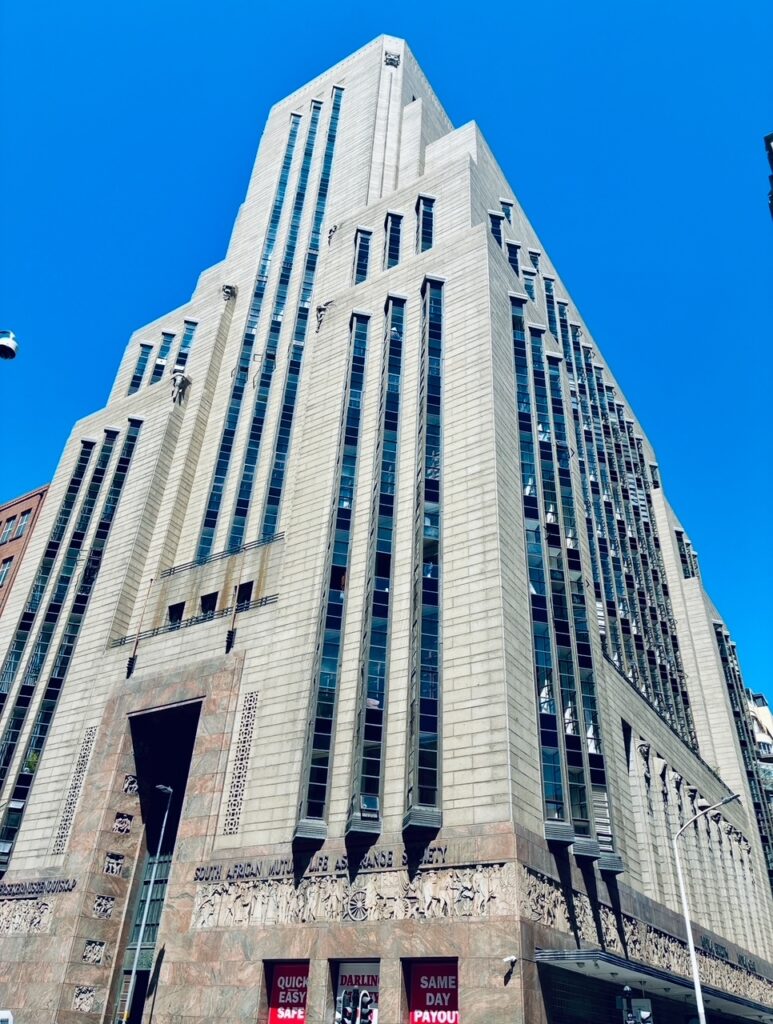 Hoch aufragendes Jugendstilgebäude mit vertikalen Fensterstreifen und dekorativem Relief am Sockel vor klarem blauem Himmel
