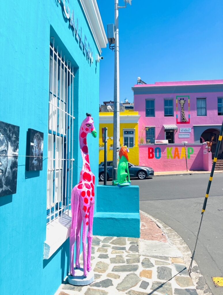 Straßenecke mit bunten Gebäuden und zwei farbigen Giraffenskulpturen auf Sockeln vor einem türkisfarbenen Haus unter klarem, strahlend blauem Himmel