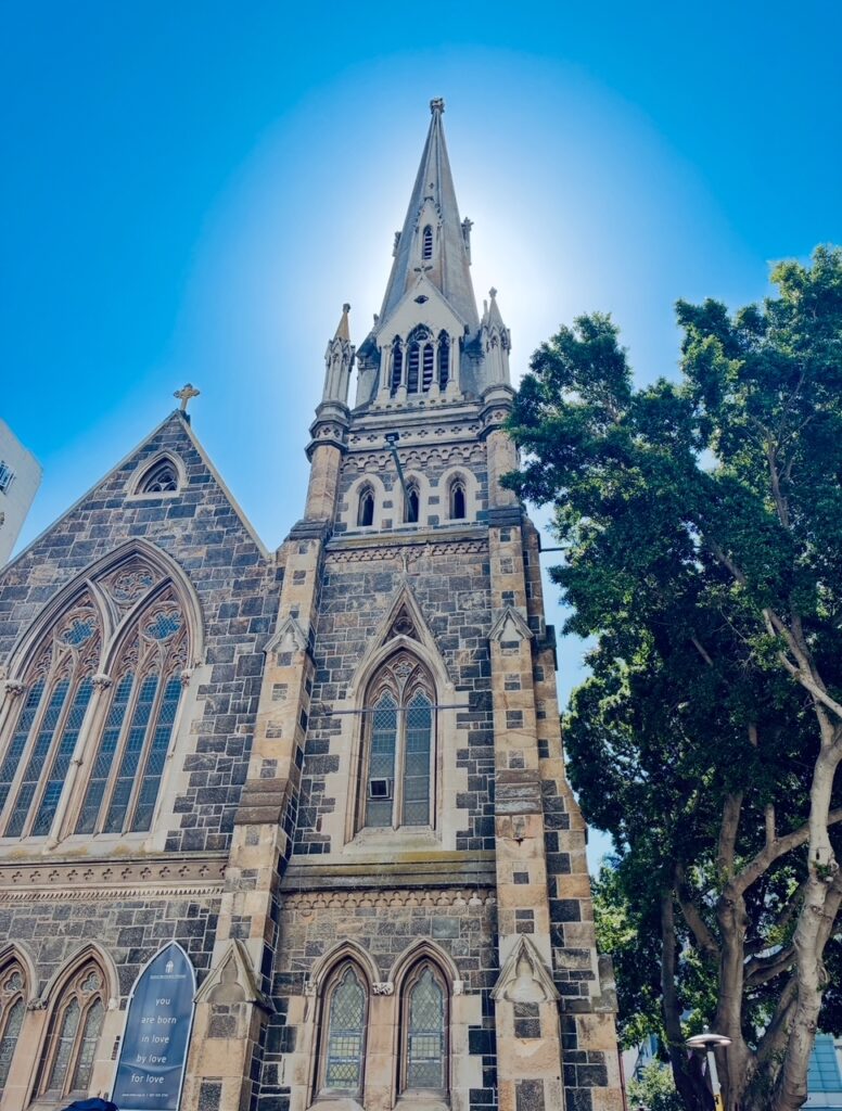 Gotische Kirche mit hohem spitzem Turm und großen Buntglasfenstern neben einem Baum unter blauem Himmel, die Sonne strahlt von hinten auf die Turmspitze