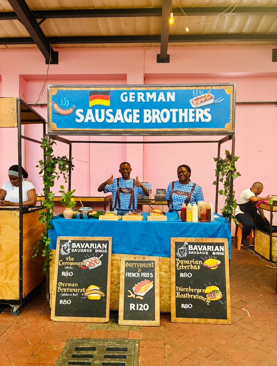 Verkaufsstand mit blauem Tisch und Schild 'GERMAN SAUSAGE BROTHERS' sowie drei Tafeln mit Speisekarte vor rosafarbener Wand und zwei lachende Schwarze Personen in karierten Hemden hinter dem Stand