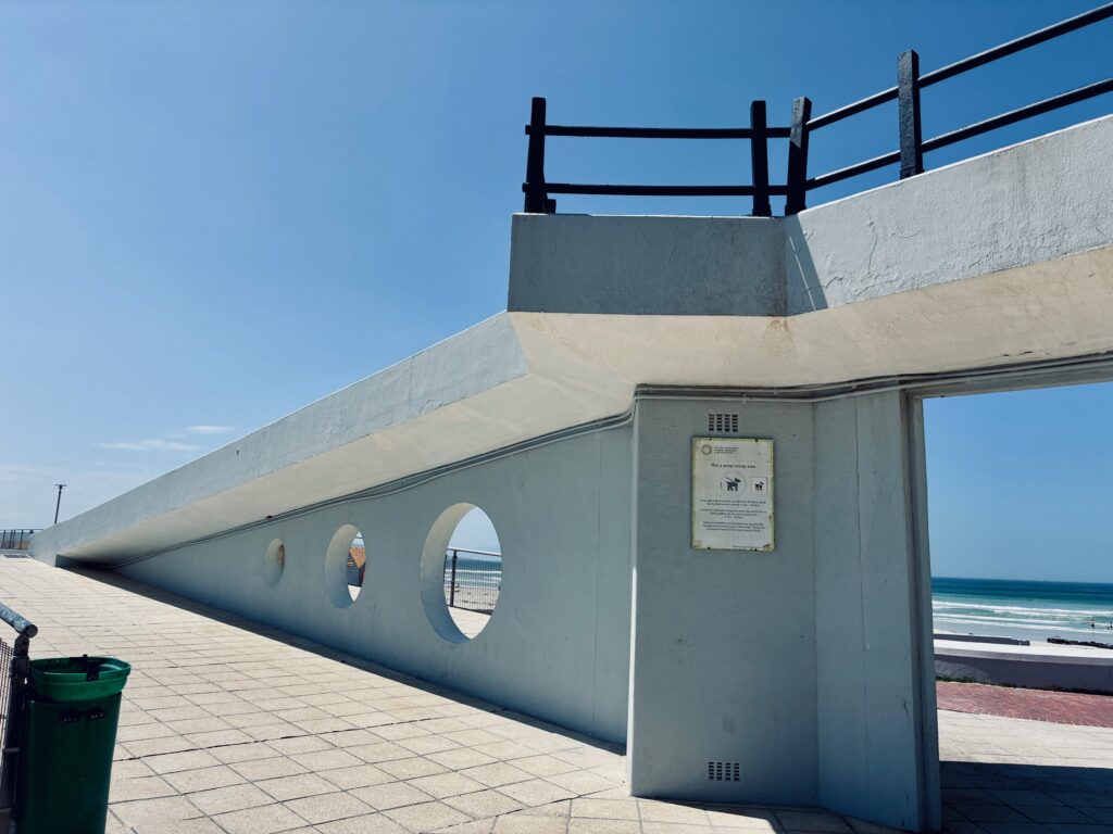 Modernistische weiße Betonmauer mit drei runden Öffnungen und Geländer am oberen Rand an einem Strand unter blauem Himmel