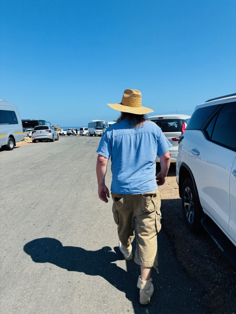 Mann mit Strohhut, blauem Hemd und beiger Hose geht auf einer Straße mit vielen geparkten Autos