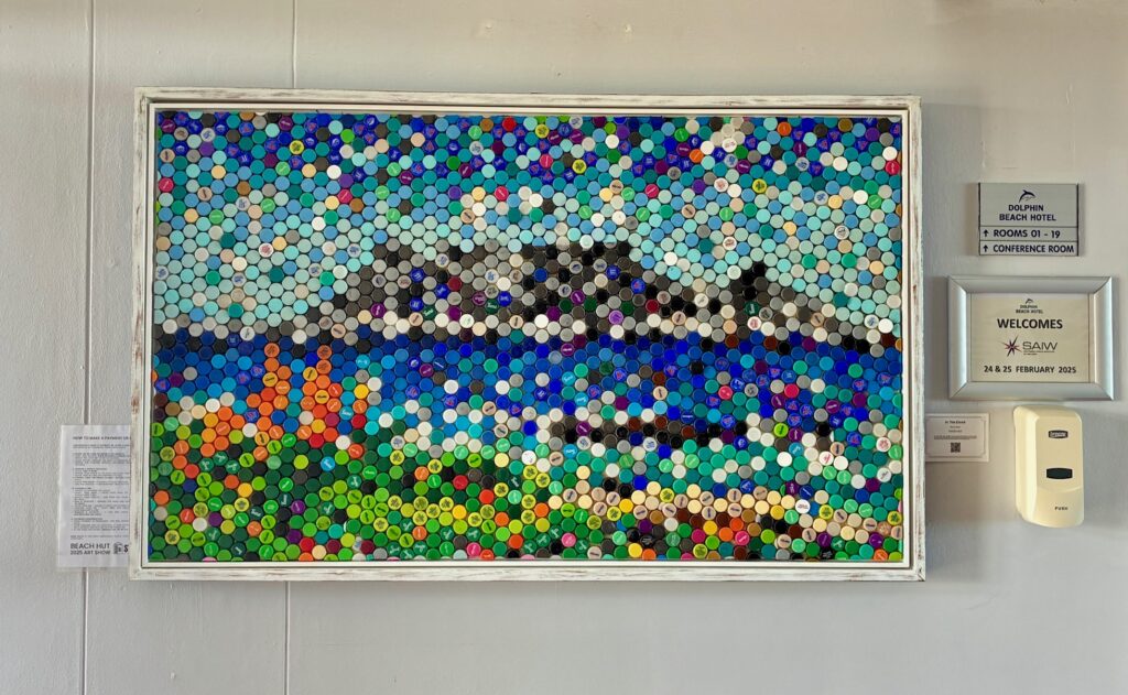 Gerahmtes Mosaik aus bunten Plastikflaschendeckeln zeigt den Tafelberg in Kapstadt und bunte Blumen im Vordergrund sowie das Meer