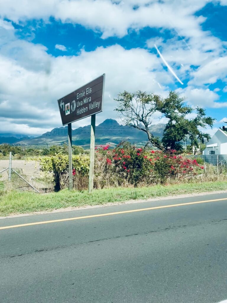 Straßenschild mit den Aufschriften 'Ernie Els', 'Uva Mira' und 'Hidden Valley' neben einer Straße mit Gebüsch und Bergen im Hintergrund