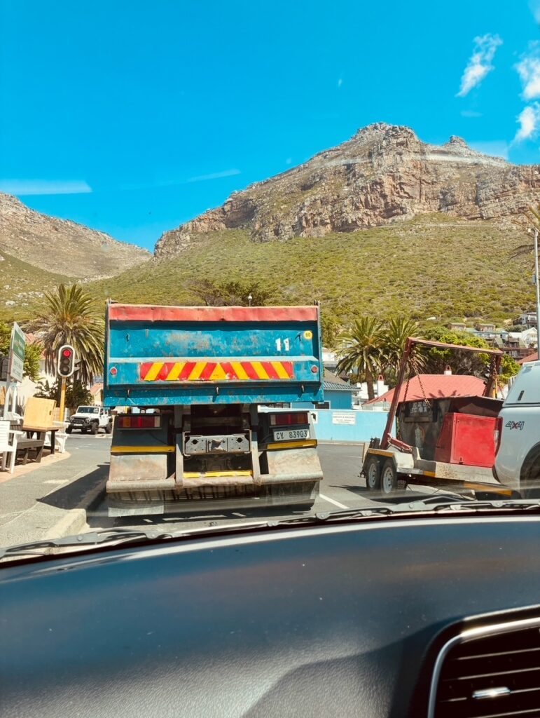 Blick aus einem Auto auf einen blauen Lastwagen mit rot-gelber Markierung vor einer Berglandschaft mit Palmen und einem roten Anhänger rechts