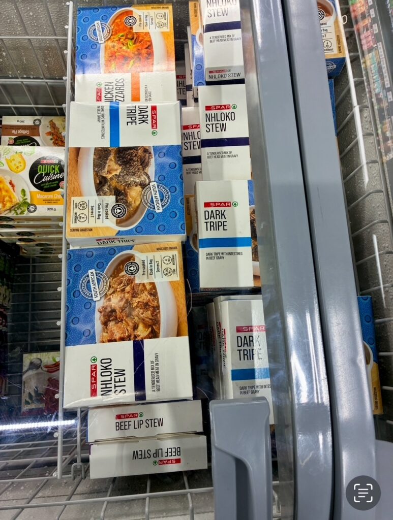 Gefrorene Fertiggerichte in Verpackungen mit den Aufschriften 'NHLOKO STEW', 'DARK TRIPE' und 'BEEF LIP STEW' in einem Supermarkt-Kühlregal