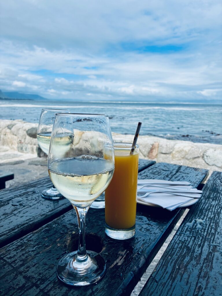 Zwei Gläser mit Weisswein und ein Glas Orangensaft auf einem Holztisch vor dem Meer unter blauem Himmel mit Wolken