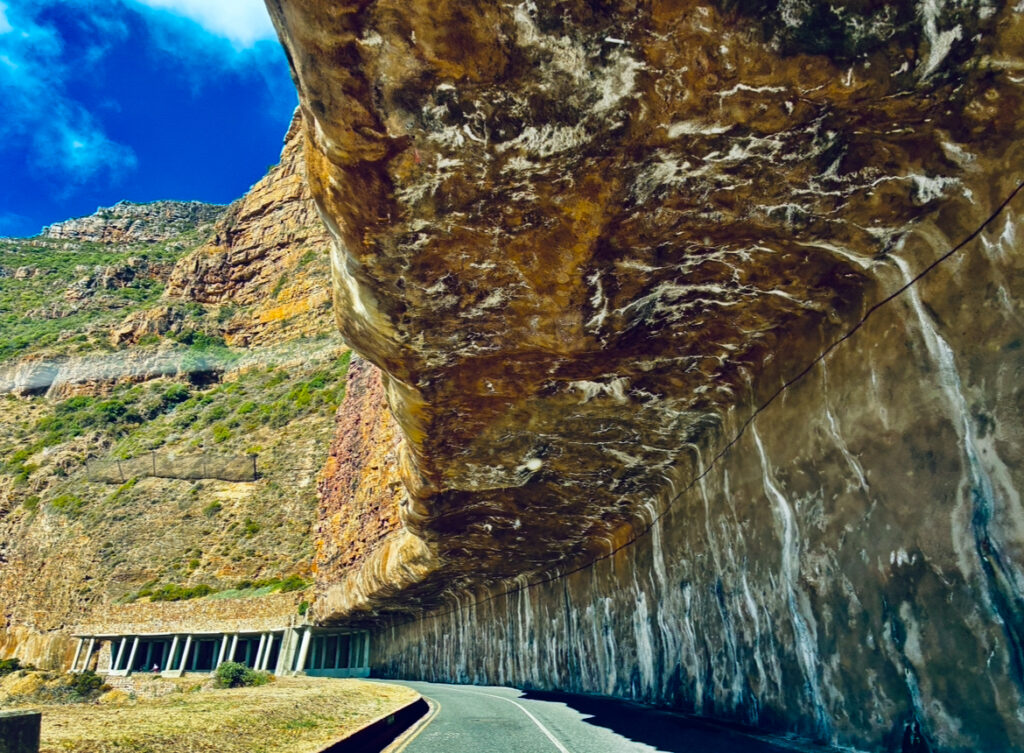 Straße führt unter einem großen, überhängenden Felsen mit sichtbaren Gesteinsstrukturen und einem blauen Himmel im Hintergrund