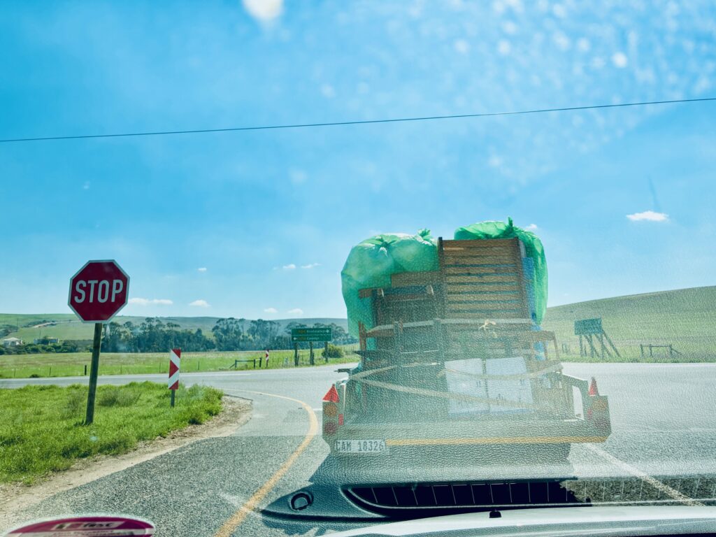 Blick durch eine Windschutzscheibe auf einen mit grünem Netz beladenen Anhänger mit Möbeln vor einem Stoppschild an einer Landstraße unter blauem Himmel