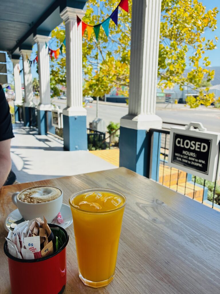 Glas mit Orangensaft und Tasse Kaffee auf Holztisch vor Veranda mit weißen Säulen und Tafel mit Öffnungszeiten