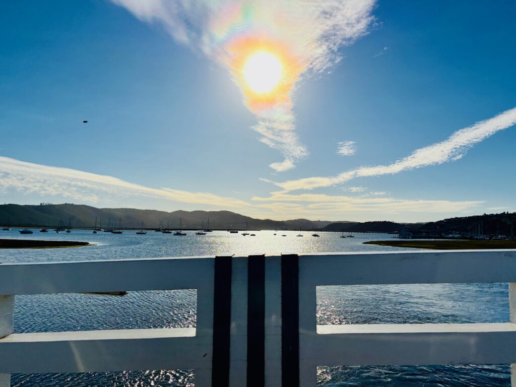 Blick über eine Wasserfläche mit mehreren Booten, im Hintergrund Hügel und ein strahlender Sonnenball am Himmel über einem weißen Geländer mit drei schwarzen Streifen