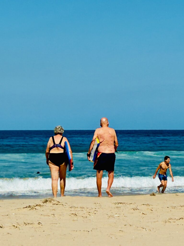 Zwei ältere Personen mit Schwimmbrettern gehen vom Sandstrand ins Meer, ein Junge läuft am Wasser entlang