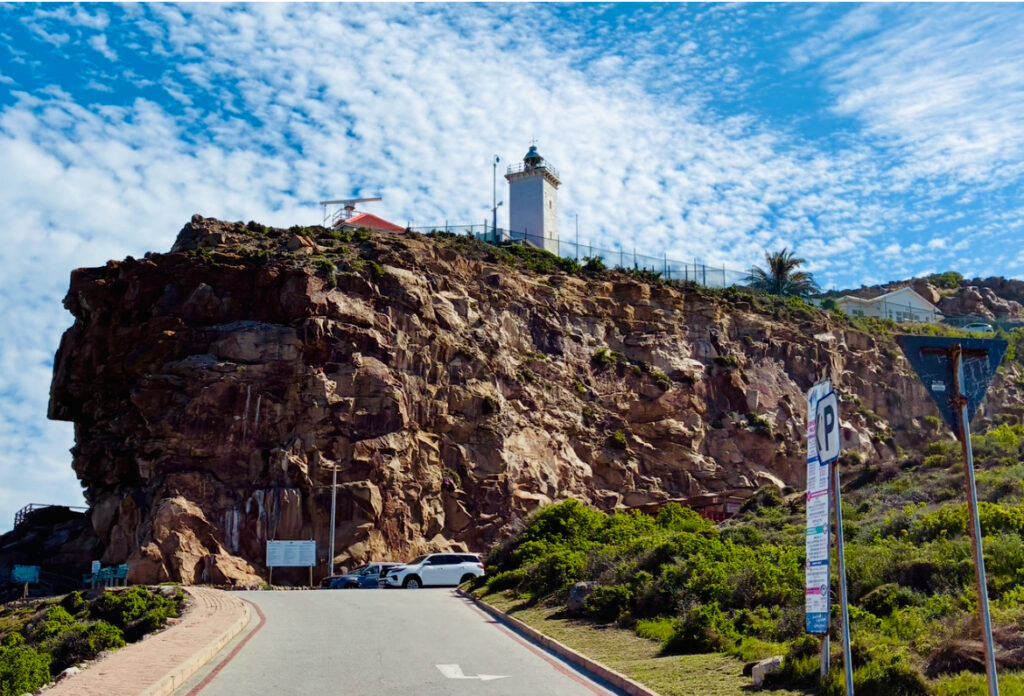 Felsklippe mit Leuchtturm und Gebäuden oben, davor Straße mit parkenden Autos und Verkehrsschildern
