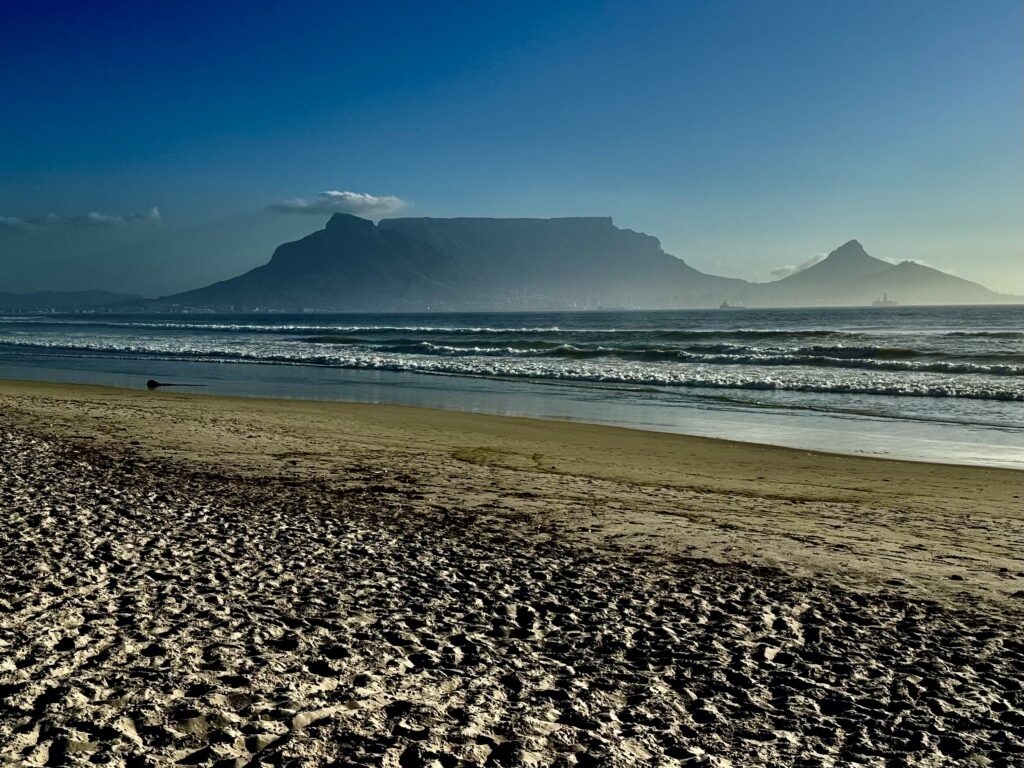 Strand mit Wellen im Vordergrund und flachem Tafelberg sowie spitzem Berg im Hintergrund unter blauem Himmel