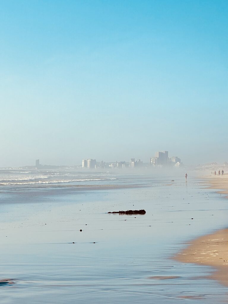Strand mit nassem Sand, im Hintergrund verschwommene, an eine Fata Morgana erinnernde Gebäude und einzelne Personen am Wasser