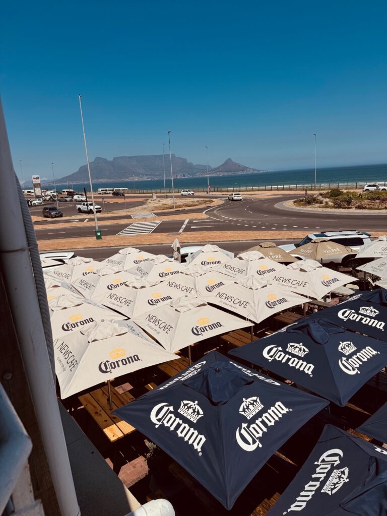 Blick auf mehrere Sonnenschirme an einem Außenbereich nahe einer Küstenstraße und Meer mit dem Tafelberg im Hintergrund