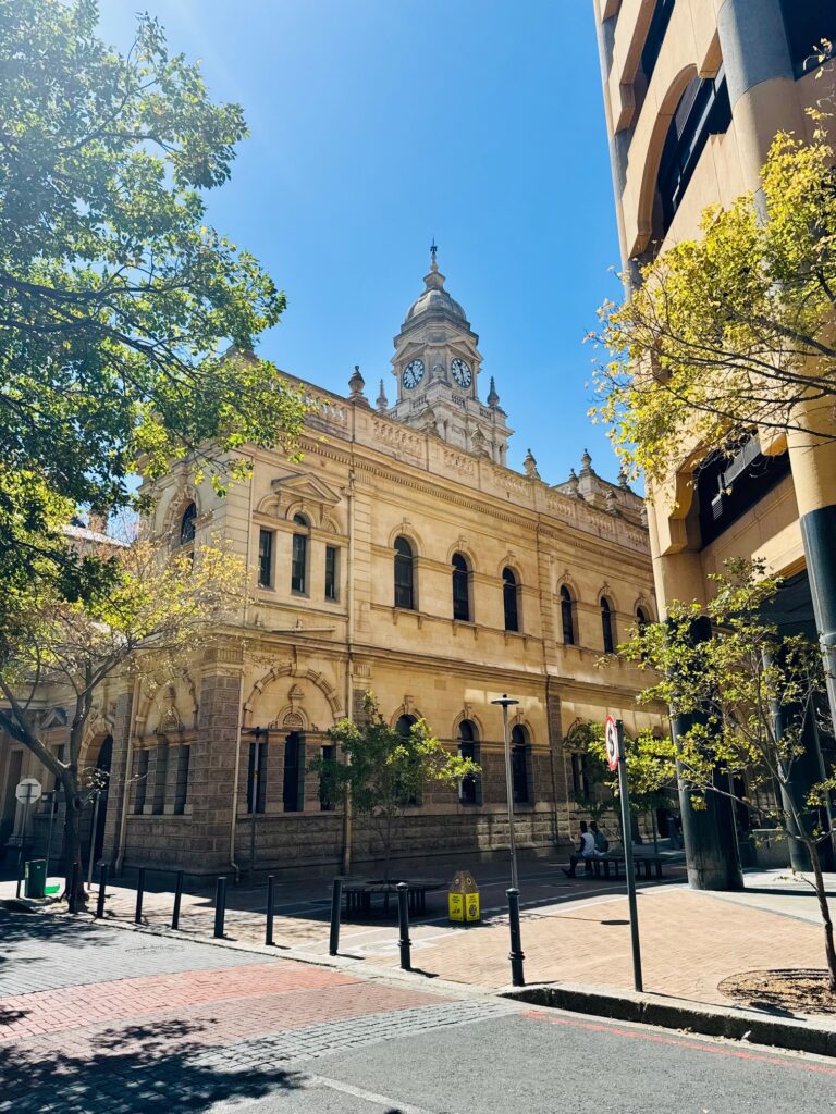 Historisches Gebäude mit Uhrenturm an einer sonnigen Straßenecke, umgeben von Bäumen und moderner Architektur