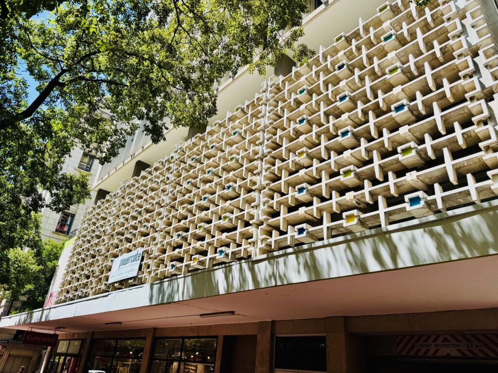 Fassade eines Gebäudes mit einem Muster aus rechteckigen Betonblöcken und kleinen farbigen Elementen im brutalistischen Stil, darüber Baumzweige mit grünen Blättern