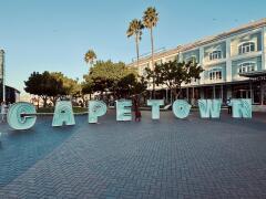 Große Buchstaben bilden das Wort 'CAPETOWN' auf einem gepflasterten Platz vor einem mehrstöckigen Gebäude mit Palmen im Hintergrund
