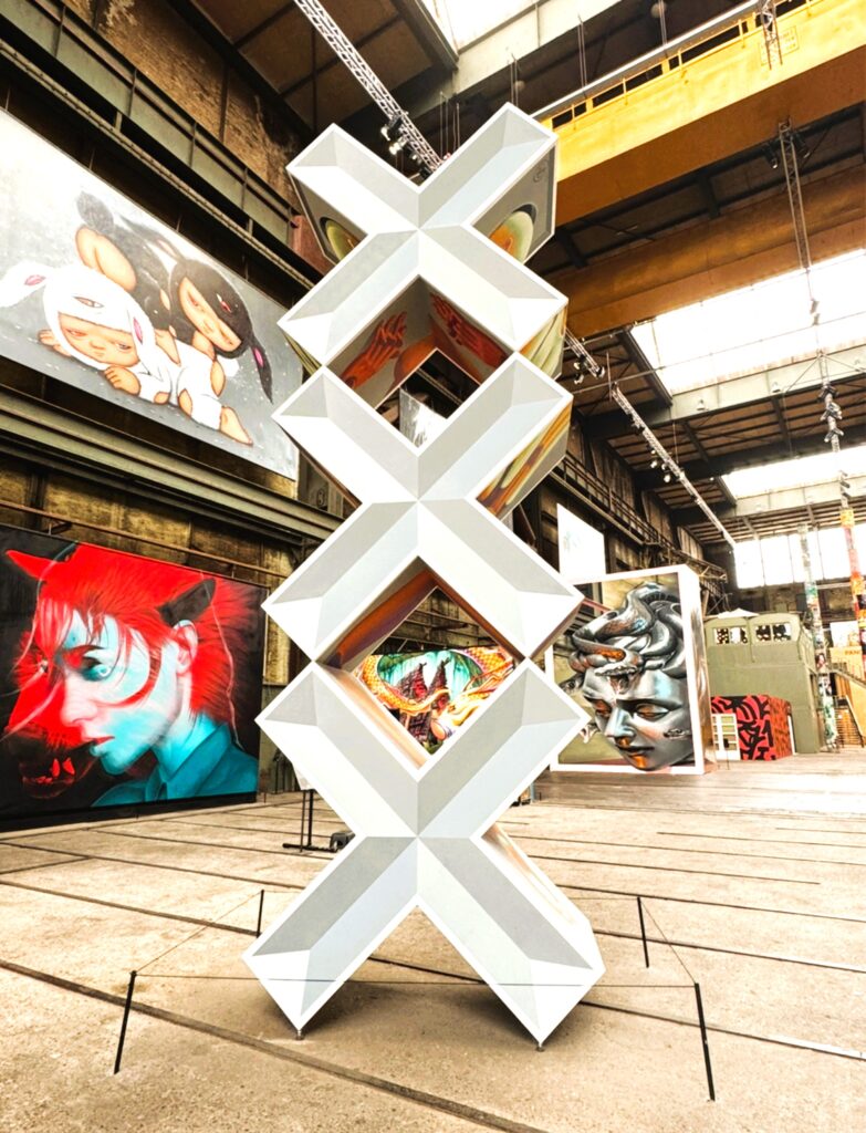 Fabrikhalle mit zahlreichen Street-Art-Kunstwerken, im Vordergrund eine Skulptur mit drei aufeinanderstehenden X-förmigen Elementen