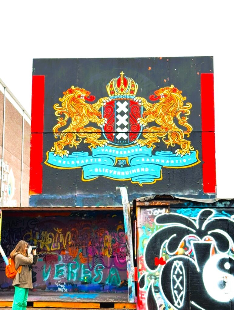 Gebäudemauer mit farbenfrohen Graffiti und Gemälden, eines zeigt das Stadtwappen von Amsterdam mit zwei Löwen, die ein Wappen mit drei aufeinander stehenden weißen Kreuzen halten