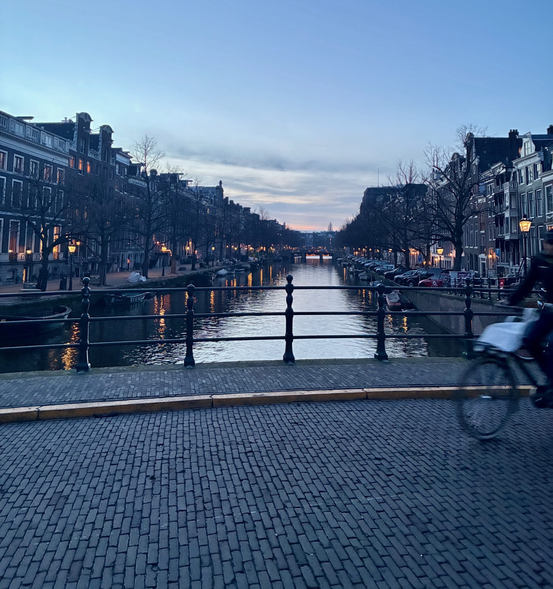 Blick auf einen Kanal in Amsterdam bei Dämmerung mit Häusern beidseitig und einer Person auf einem Fahrrad im Vordergrund