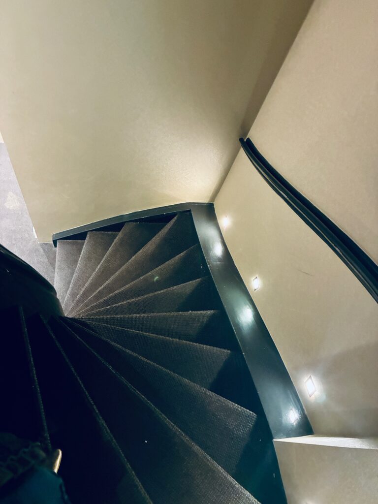 Steile Wendeltreppe mit dunklem Teppich und Handlauf an der Wand, mit Lichtern beleuchtet