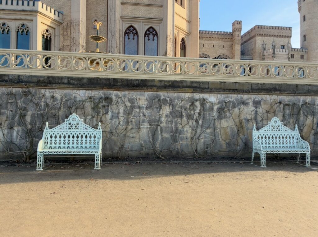 Zwei verschnörkelte Metallbänke vor einer zieselierten Steinmauer mit einem Schloss im Hintergrund