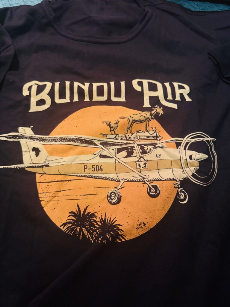 Dunkles T-Shirt mit Vintage Aufdruck "Bundu Air", das eine historische Propellermaschine vor einem gelben Mond zeigt, mit auf dem Dach des Flugzeugs stehender Ziege und Hahn