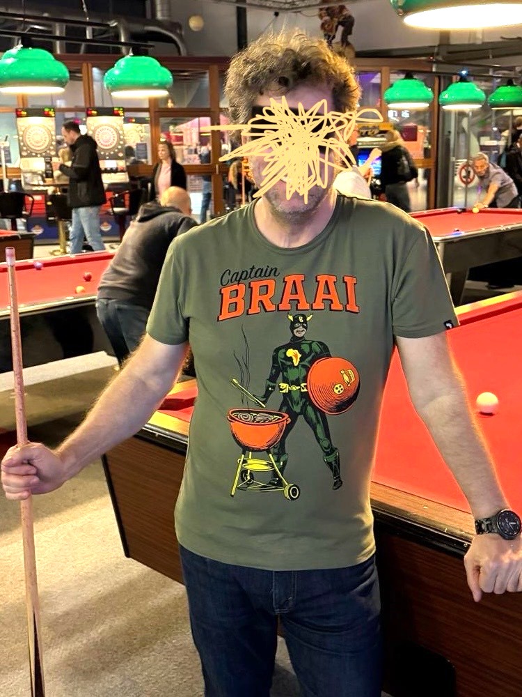 Mann mit T-Shirt mit Aufdruck "Captain BRAAI" und Comicfigur mit Grill und Grilldeckel lehnt an Billardtisch und hält Billardqueue, Gesicht des Mannes ist unkenntlich gemacht
