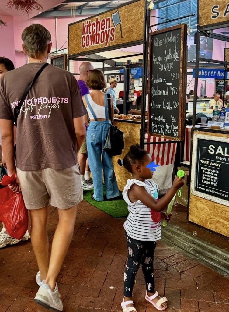 Markthalle mit Ständen und einem Schild 'kitchen cowboys' sowie einer Tafel mit Speisenangebot, davor Menschen, darunter ein Mädchen mit weißen Sandalen und gestreiftem Shirt